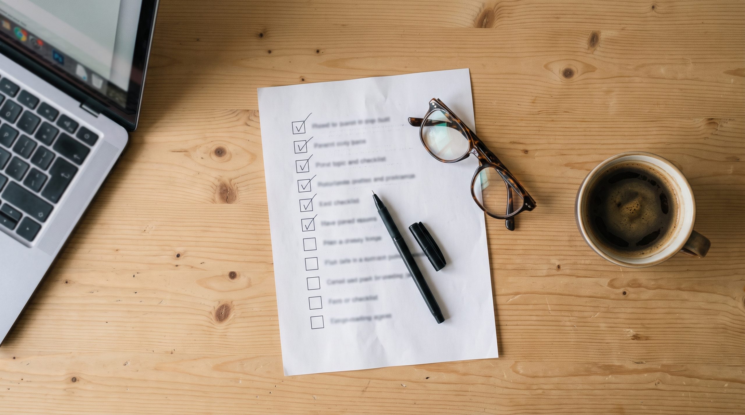 Printed checklist with tick marks next to a laptop and pen on a desk