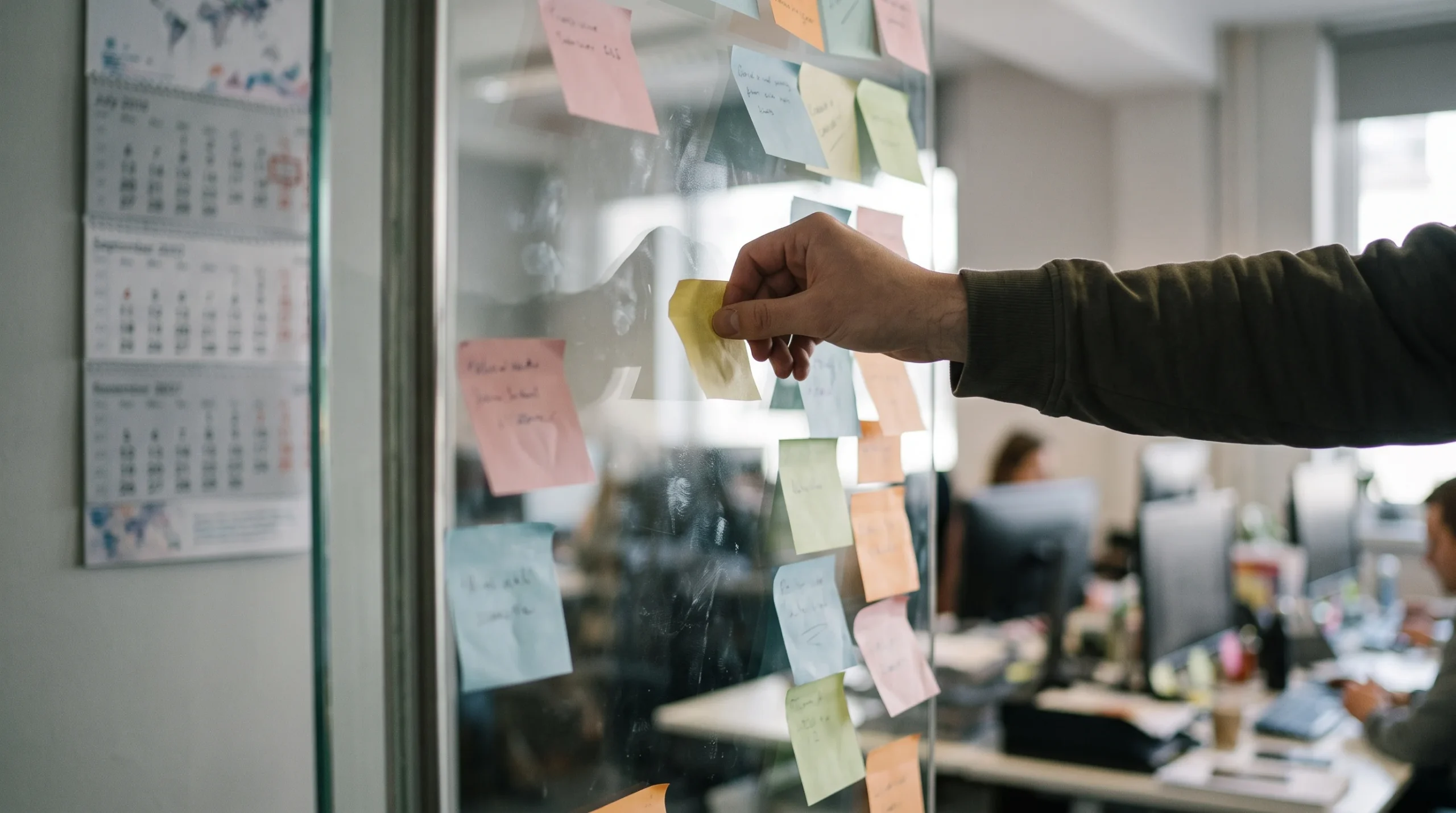 Hand placing a sticky note on a glass planning board next to a wall calendar